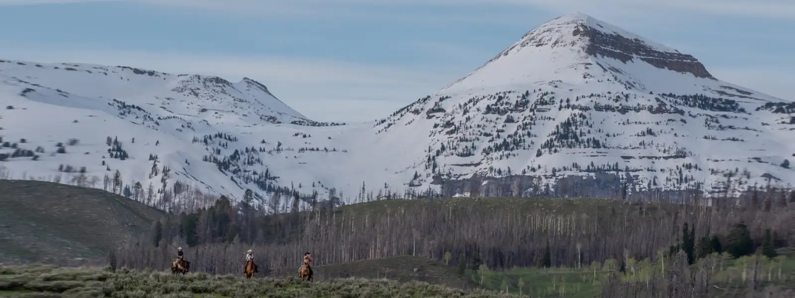 Three people riding on horseback with two Gros Ventre mountain peaks in the background.